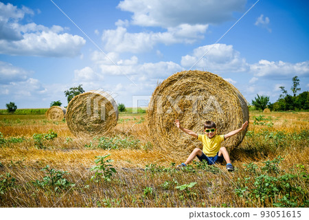 Little boy sits on a round haystack relaxed with hands up. Field 93051615