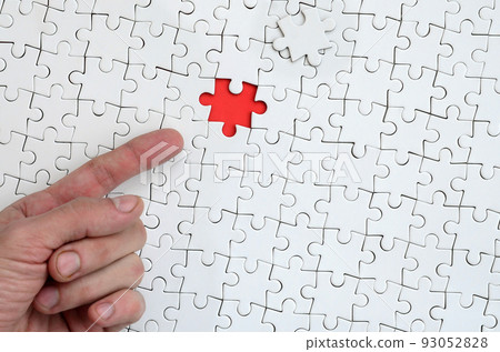 The texture of a white jigsaw puzzle in the assembled state with one missing element, forming a red space, pointed to by the finger of the male hand 93052828