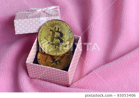 The golden bitcoin lies in a small pink gift box with a small bow on a blanket made of soft and fluffy light pink fleece fabric with a large number of relief folds 93053406