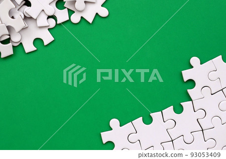 Fragment of a folded white jigsaw puzzle and a pile of uncombed puzzle elements against the background of a green surface. Texture photo with space for text 93053409