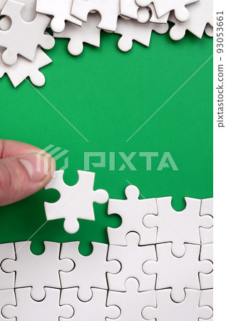 The hand folds a white jigsaw puzzle and a pile of uncombed puzzle pieces lies against the background of the green surface. Texture photo with space for text The hand folds a white jigsaw puzzle and a pile of uncombed puzzle pieces lies against the background of the green surface. Texture photo with space for text 93053661