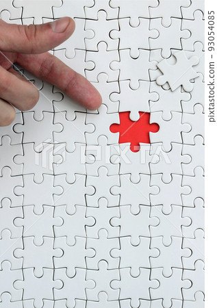 The texture of a white jigsaw puzzle in the assembled state with one missing element, forming a red space, pointed to by the finger of the male hand 93054085