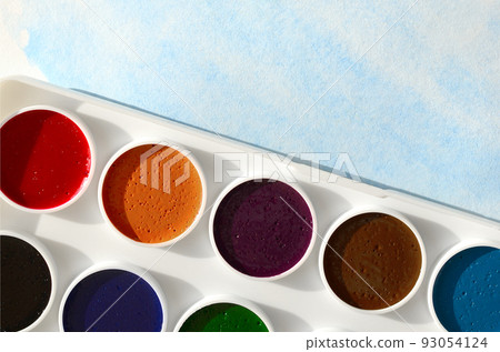 A new set of watercolors lies on a sheet of paper, which shows an abstract watercolor drawing in the form of blue strokes. The concept of amateur painting among teenagers A new set of watercolors lies on a sheet of paper, which shows an abstract watercolor drawing in the form of blue strokes. The concept of amateur painting among teenagers 93054124
