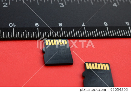 Two small micro SD memory cards lie on a red background next to a black ruler. A small and compact data and information store Two small micro SD memory cards lie on a red background next to a black ruler. A small and compact data and information store 93054203