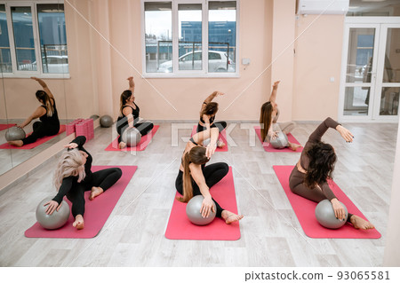 A group of six athletic women doing pilates or yoga on pink mats in front of a window in a beige loft studio interior. Teamwork, good mood and healthy lifestyle concept. A group of six athletic women doing pilates or yoga on pink mats in front of a window in a beige loft studio interior. Teamwork, good mood and healthy lifestyle concept. 93065581