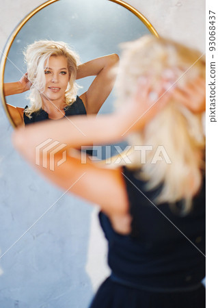 an attractive blonde in a black dress looks at the reflection in the mirror  93068437