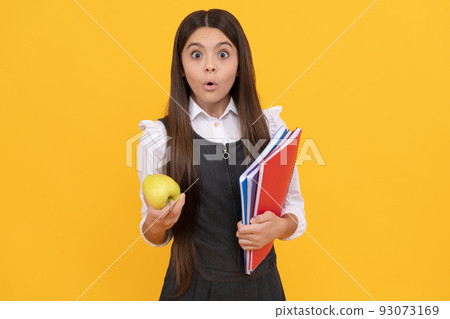 Surprised girl child hold apple and school textbooks yellow background, homework 93073169