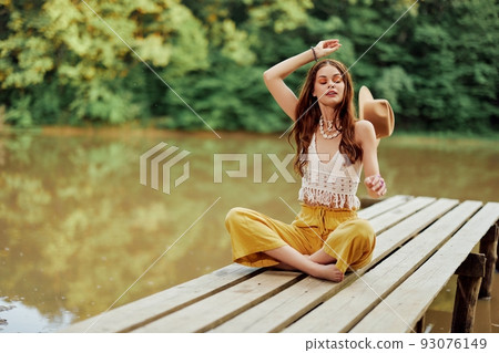 A young hippie woman sits on a lake bridge wearing stylish eco clothes and smiling A young hippie woman sits on a lake bridge wearing stylish eco clothes and smiling 93076149