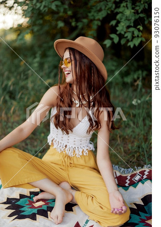 A young hippie woman in eco-clothing has fun laughing and cringing sitting on a plaid outside in the fall with a hat and yellow sunglasses. Travel lifestyle A young hippie woman in eco-clothing has fun laughing and cringing sitting on a plaid outside in the fall with a hat and yellow sunglasses. Travel lifestyle 93076150