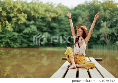 Hippie eco-activist woman traveler sits on a bridge by a lake with her arms outstretched with a hat and smiling sincerely Hippie eco-activist woman traveler sits on a bridge by a lake with her arms outstretched with a hat and smiling sincerely 93076152