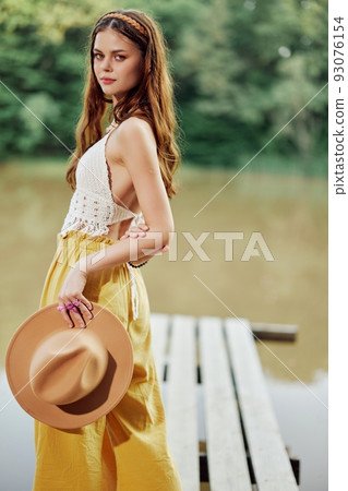 A young woman smiling in an image of a hippie and eco-dress dancing in nature by the lake wearing a hat and yellow pants in the summer sunset A young woman smiling in an image of a hippie and eco-dress dancing in nature by the lake wearing a hat and yellow pants in the summer sunset 93076154