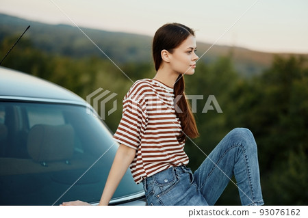 A young woman traveler sits on the trunk of her car and watches the sunset in nature. The concept of freedom and travel as a lifestyle A young woman traveler sits on the trunk of her car and watches the sunset in nature. The concept of freedom and travel as a lifestyle 93076162