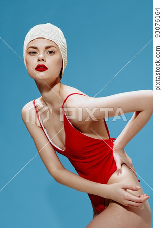 A spectacular flexible young female swimmer in a red swimsuit cap bends posing on a blue background in the studio A spectacular flexible young female swimmer in a red swimsuit cap bends posing on a blue background in the studio 93076164