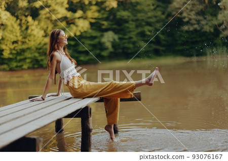A young happy hippie woman sitting on the lake shore on a bridge in eco clothing made of natural materials in harmony with nature in the fall A young happy hippie woman sitting on the lake shore on a bridge in eco clothing made of natural materials in harmony with nature in the fall 93076167