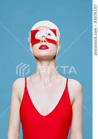 A bright red-lipped lady in a red swimsuit glasses swimming cap raises her head up posing isolated on a blue background in the studio 93076187