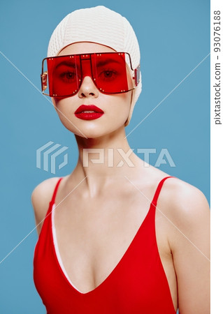 A lady in a red swimsuit, swimming cap, glasses looks at the camera on a blue background. Fashion concept for brands 93076188