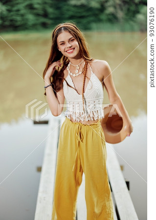 A young woman in a hippie look and eco-dress dancing outdoors by the lake wearing a hat and yellow pants in the summer sunset 93076190