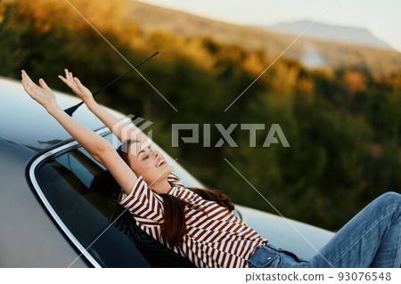 The woman driver stopped on the road and lay down on the car to rest and look at the beautiful landscape in a striped T-shirt and jeans. Complicated journey to nature The woman driver stopped on the road and lay down on the car to rest and look at the beautiful landscape in a striped T-shirt and jeans. Complicated journey to nature 93076548