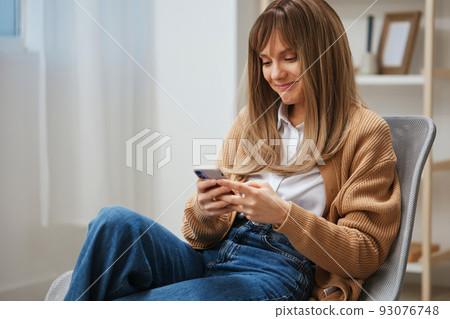 Happy cute blonde student lady in warm sweater chatting with boyfriend use phone sitting in armchair at home interior. Pause from work, take a break, social media in free time concept. Copy space 93076748