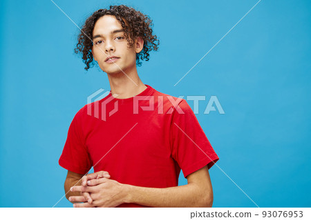 Attractive cute young curly tanned Caucasian guy in basic red t-shirt looks at camera posing isolated on over blue background. Lifestyle and Emotions concept. Good offer with copy space for ad Attractive cute young curly tanned Caucasian guy in basic red t-shirt looks at camera posing isolated on over blue background. Lifestyle and Emotions concept. Good offer with copy space for ad 93076953