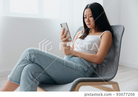 Focused thoughtful young Asian woman read shock information using phone sit on chair at home interior living room. Distance communication, Remote work, Social media concept. Cool offer Banner Mockup Focused thoughtful young Asian woman read shock information using phone sit on chair at home interior living room. Distance communication, Remote work, Social media concept. Cool offer Banner Mockup 93077208