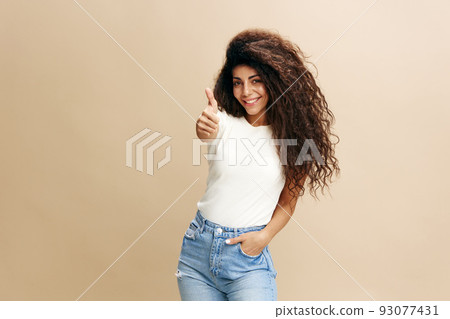 Enjoyed stylish curly Latin female in brown hat, smiling at camera, showing thumb up, saying Yeah great idea, isolated beige background. Copy space clothing fashion brands, free place for your ad. Enjoyed stylish curly Latin female in brown hat, smiling at camera, showing thumb up, saying Yeah great idea, isolated beige background. Copy space clothing fashion brands, free place for your ad. 93077431