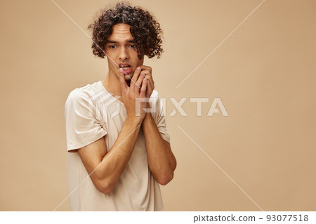Confused awesome tanned curly man in basic t-shirt touches cheeks posing isolated on over beige pastel background. Fashion New Collection offer. People and Emotions concept. Free place for ad 93077518