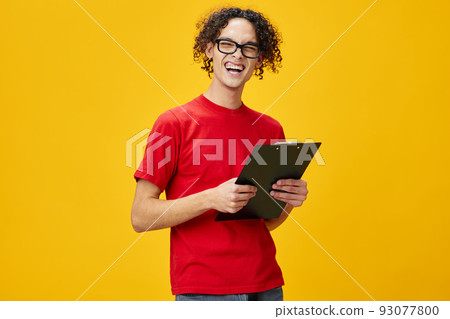 Emotional myopic young student man in red t-shirt funny eyewear holds tablet folder with study notes posing isolated on over yellow studio background. Free place for ad. Education College concept Emotional myopic young student man in red t-shirt funny eyewear holds tablet folder with study notes posing isolated on over yellow studio background. Free place for ad. Education College concept 93077800
