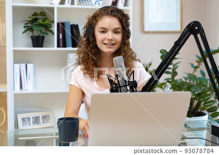 Young curly female recording podcast from home and talking into a microphone 93078784