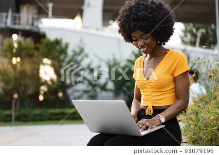 Young african american woman working on laptop sitting on the bench at the city Young african american woman working on laptop sitting on the bench at the city 93079196