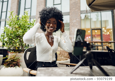 Portrait of smiling woman with afro hairstyle on video call sitting in cafe Portrait of smiling woman with afro hairstyle on video call sitting in cafe 93079199