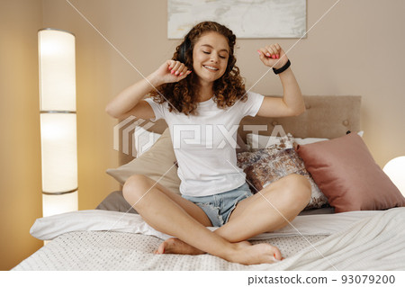 Young happy woman listening music in headphones with smartphone in bed Young happy woman listening music in headphones with smartphone in bed 93079200