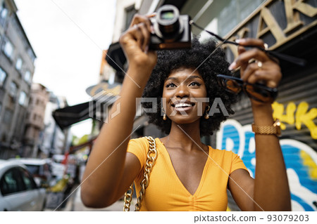 Smiling African american woman using professional camera at a street Smiling African american woman using professional camera at a street 93079203