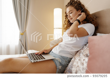 Portrait of a young curly woman using laptop in bed at home Portrait of a young curly woman using laptop in bed at home 93079205