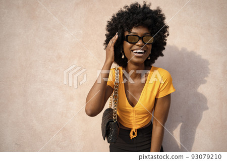 Happy African American young woman with afro hair wearing stylish sunglasses posing against beige wall background Happy African American young woman with afro hair wearing stylish sunglasses posing against beige wall background 93079210