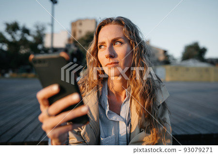 Young casual woman in coat sitting on bench on sea embankment using smartphone 93079271