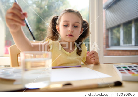 Charming creative European schoolgirl, talented little artist, dips a paintbrush into a glass of water while painting with watercolors. Craetive art class. Kids entertainment. Drawing. Painting. 93080869