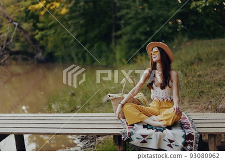 Woman in hippie eco dress sits in nature by the lake on a bridge relaxing with a basket of food and admiring the scenery watching the sunset 93080962