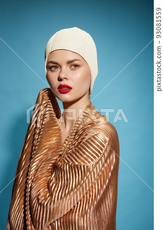 Goldfish concept. A girl with red lips in a gold scarf and a swimming cap looks away on a blue background Goldfish concept. A girl with red lips in a gold scarf and a swimming cap looks away on a blue background 93081559
