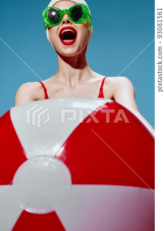 A screaming cheerful unusual lady in a swimming cap pulls a red white ball into the camera on a blue background. Time to go to the sea 93081561