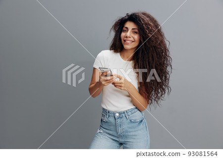 Pretty young african american girl in white t-shirt posing isolated on gray background, studio portrait. People lifestyle concept. Mock up copy space. Using mobile phone, typing message, social media Pretty young african american girl in white t-shirt posing isolated on gray background, studio portrait. People lifestyle concept. Mock up copy space. Using mobile phone, typing message, social media 93081564