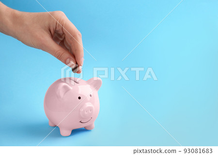 Woman putting coin into piggy bank on light blue background, closeup. Space for text Woman putting coin into piggy bank on light blue background, closeup. Space for text 93081683