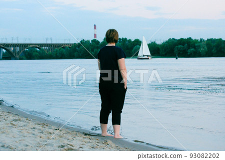 A woman with a short haircut stands on the sandy shore of the sea, river and looks out into the distance on a white sailboat. Loneliness, sadness, brooding concept. Traveling solo. Urban city scene. 93082202