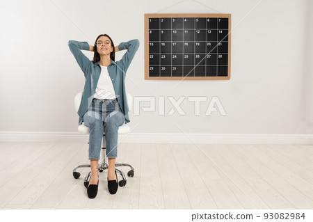 Beautiful young woman sitting near board calendar indoors 93082984