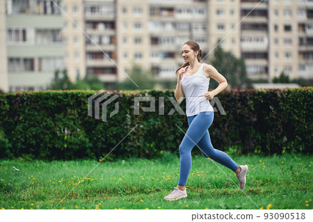 Woman in white t-shirt and leggings takes morning jog in residential area of city next to high-rise apartment building. Woman in white t-shirt and leggings takes morning jog in residential area of city next to high-rise apartment building. 93090518