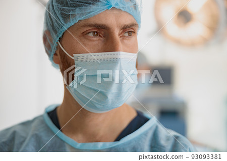 Close up of professional doctor in mask standing in operating room before surgery and looking away Close up of professional doctor in mask standing in operating room before surgery and looking away 93093381