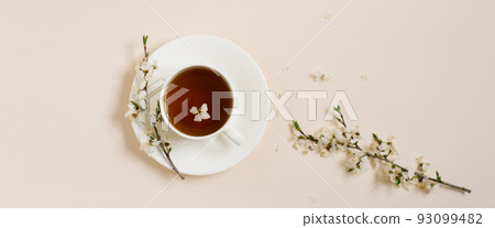 White porcelain cup with black tea and marshmallows. Branches of a blossoming apple tree lie on a beige background. Spring concept. Baner White porcelain cup with black tea and marshmallows. Branches of a blossoming apple tree lie on a beige background. Spring concept. Baner 93099482
