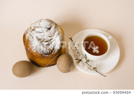 A traditional Easter cake sprinkled with powdered sugar, colored beige eggs and a cup of black tea in a white mug and a sprig of apple blossom. Holiday greeting card A traditional Easter cake sprinkled with powdered sugar, colored beige eggs and a cup of black tea in a white mug and a sprig of apple blossom. Holiday greeting card 93099484