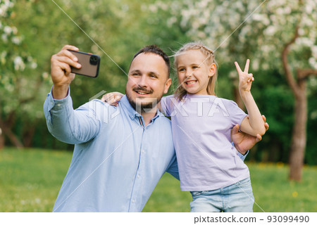 Dad and his six-year-old daughter take selfies on the phone, smiling and having fun on camera in the park in summer or spring Dad and his six-year-old daughter take selfies on the phone, smiling and having fun on camera in the park in summer or spring 93099490