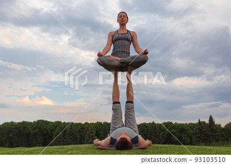 Man lying on grass and balancing woman in his feet. Young couple doing acro yoga in park Man lying on grass and balancing woman in his feet. Young couple doing acro yoga in park 93103510